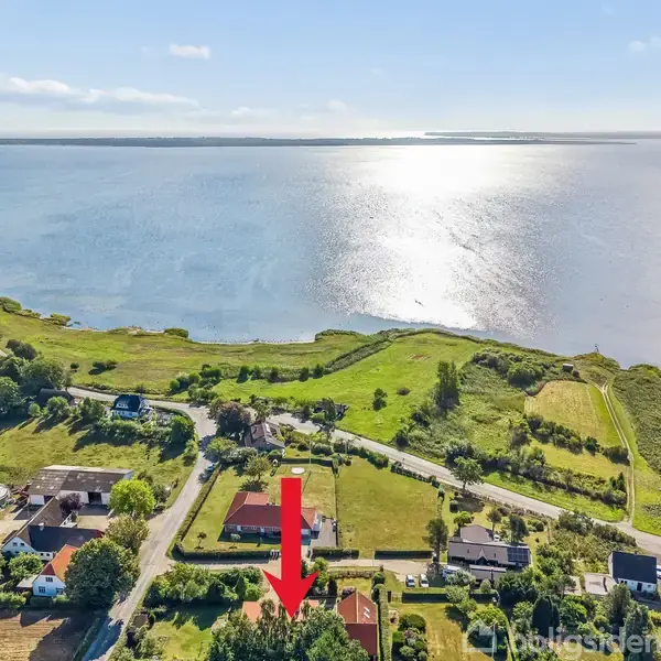 Rødt hus markeret med pil står blandt grønne områder nær en sø. Solen reflekterer i vandet, omgivet af flere huse og veje.
