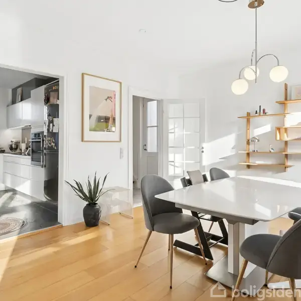Et elegant spisebord med grå stole står i et lyst rum med parketgulv. Til venstre ses et moderne køkken med hvide skabe. Hylder med dekorationer hænger på væggen.