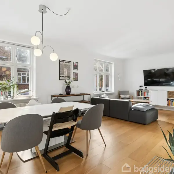 Et moderne stueområde med en hvid spisebord, omgivet af stole, og en grå sofa foran en vægmonteret fladskærms-TV. Trægulve og lyse vægge med vinduer skaber en lys atmosfære.