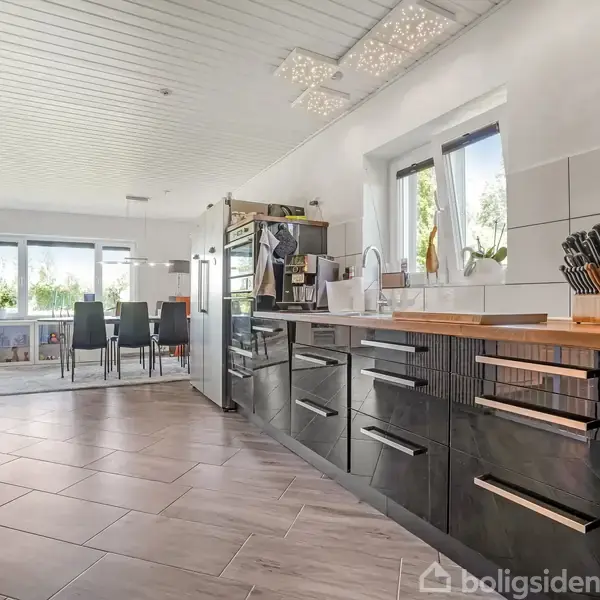 Et moderne køkken med sorte skabe og træbordplade. Rummet har hvide vægge, vinduer med udsigt til planter, og der er spiseplads i baggrunden med sorte spisestole ved vinduet.