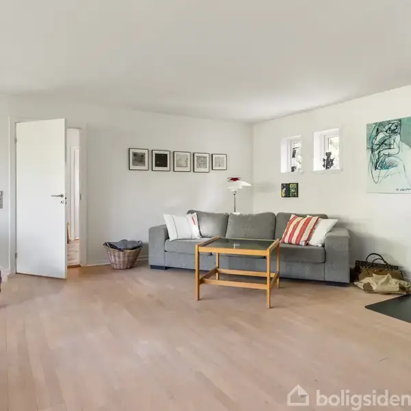 En stue med sofa og puder ved siden af en brændeovn. På væggen hænger kunstværker. Der er et træbord i midten, og en stol til venstre.