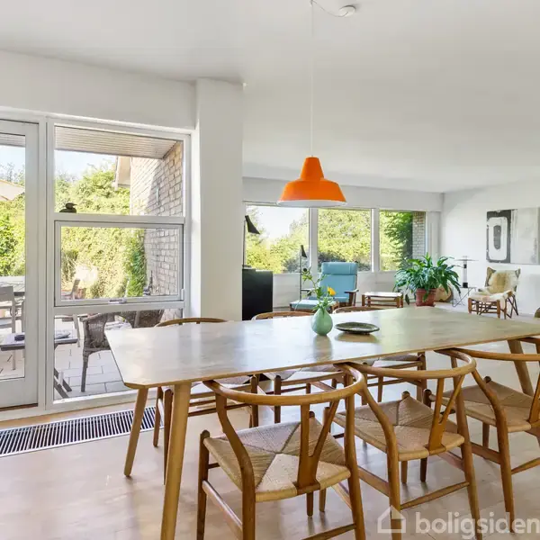 Spisebord med stole står i en lys stue med store vinduer. En orange loftslampe hænger over bordet. Udenfor er en terrasse med møbler og grønne buske.
