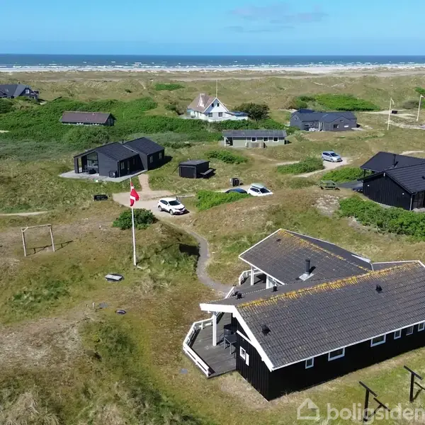 Sort hus med veranda i græslandskab, omgivet af spredte huse og køretøjer. Dannebrog vajer ved siden af hovedhuset. I baggrunden ses klitter og havet under blå himmel.
