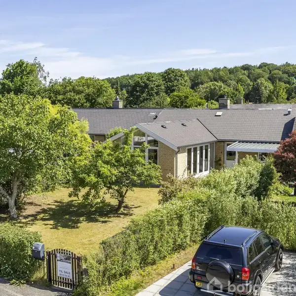 En sort bil står parkeret foran et hus med en velplejet have. Haven har grønne træer og buske. Baggrunden er en frodig skov. Skilt ved indgangen viser "Boligsiden".