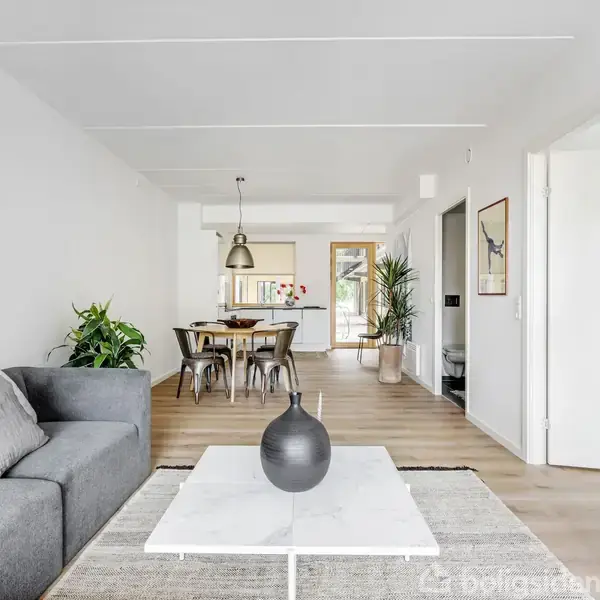 En grå sofa står ved siden af et hvidt sofabord med en sort vase. Spisebord bagved med seks stole. Rummet er lyst med trægulv og planter.