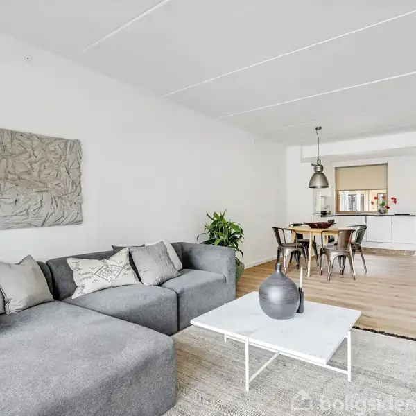 Grå hjørnesofa med puder står i en stue med lyst trægulv. Foran sofaen er et hvidt sofabord. I baggrunden ses et spisebord og åbent køkken.