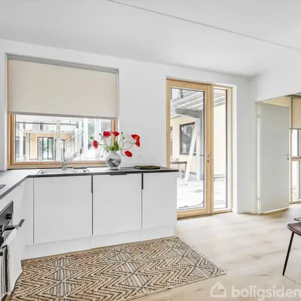 En moderne køkken med hvide skabe, en sort bordplade og en vase med røde blomster på bordet. Store vinduer og dør giver lys og udsigt til terrasse. En brun stol står ved siden af en tæppebelagt gangvej.