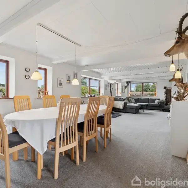 Spisebordet er dækket med hvid dug og omgivet af træstole i en lys stue. Dyreopsats hænger på væggen, og store vinduer giver udsigt til naturen udenfor.