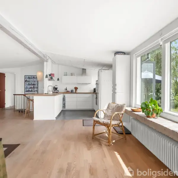 Køkken-alrum med åbent layout og trægulve. En rattan-stol står nær store vinduer, der giver udsigt til en grønt omgivelse. Køkkenet har hvide skabe og køkkenbord.