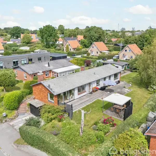En etplansvilla med gråt tag står i en frodig villakvarter med velplejede haver. Der er en terrasse med møbler, omgivet af grøn bevoksning. Blomsterbede pryder forhaven.