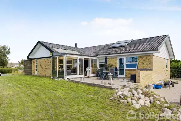 En moderne gulstensvilla med sorte tagsten står i en have med græsplæne og stenterasse. På terrassen er der havemøbler, og en glasudestue forbinder indvendige rum med udendørsområdet.