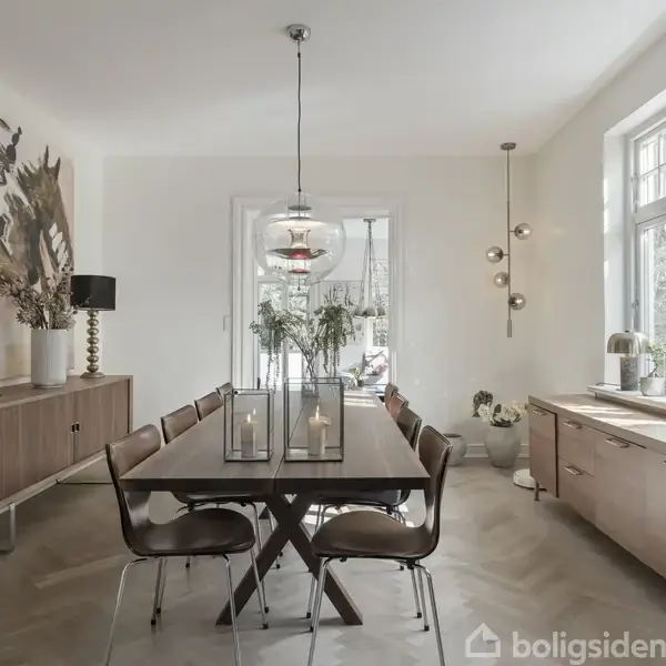 Spisebord med stole står i en moderne spisestue. Bordet er dækket med lys og planter. Omgivet af to skænke, kunst på væggen og store vinduer.