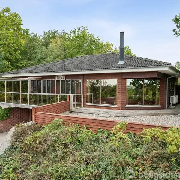 Huset står stille omgivet af tæt løvskov, med en stor glasudestue og et grusbelagt terrasseområde. Det har en mørk flisebelagt tag og brune trævægge.