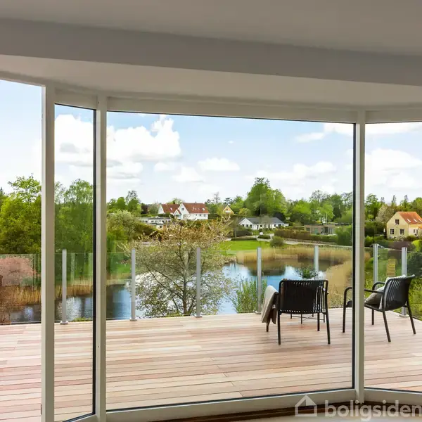 Kigger ud gennem store glasdøre mod en rummelig terrasse med to stole. Terrassen har udsigt over en sø og et grønt landskab med huse i baggrunden.