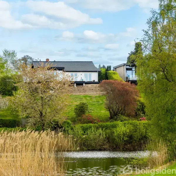 Hus står stille på bakketop med moderne arkitektur. En have med træer omkranser bygningen. I forgrunden ses en sø og siv med en advarselstrekant ved kanten.