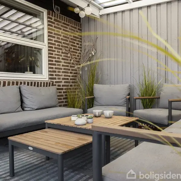 Grå sofaer med lysegrå hynder står omkring et træbord i en overdækket terrasse. Murstensvæg og gråmalede trævægge omgiver området, dekoreret med planter og farverige vægpynt.