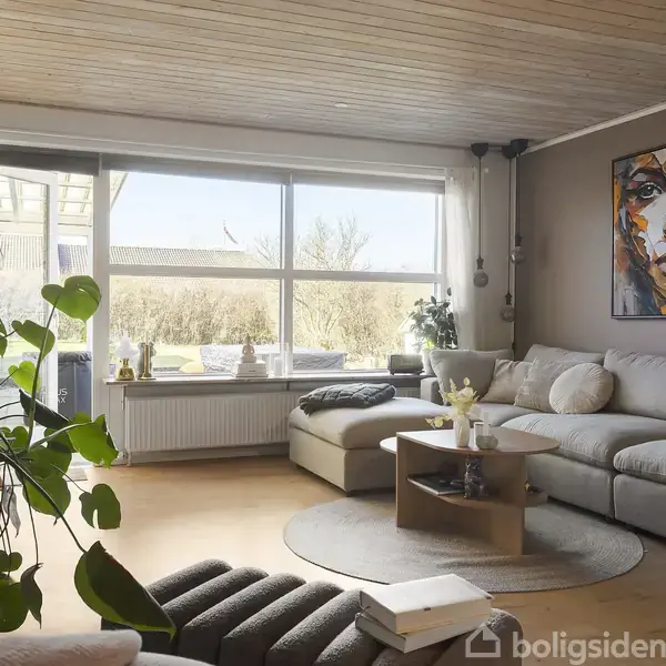 Sofa står i stuen, hvor den er omkranset af grønne planter på gulvet. Et stort, abstrakt portræt hænger på væggen. Store vinduer viser en lys have udenfor.