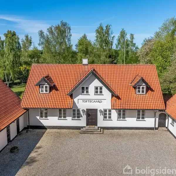 Hvid bygning med rødt tegltag, skilt med teksten "TOTTEGAARD" ovenfor døren, omgiven af grusbelagt gårdsplads. Omgivet af grønne træer og blå himmel.