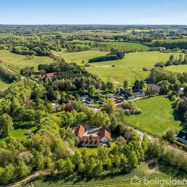En række gårde omgivet af frodige grønne marker og skove ligger spredt i et landligt område under en klar blå himmel.