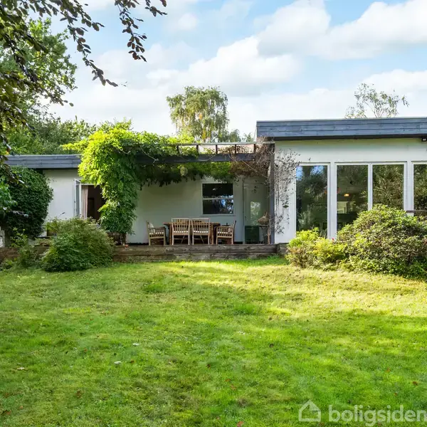Hus med hvid facade og store vinduer står stille. En terrasse med havemøbler er dækket af en pergola med grønne planter. Græsplæne og buske omringer huset.
