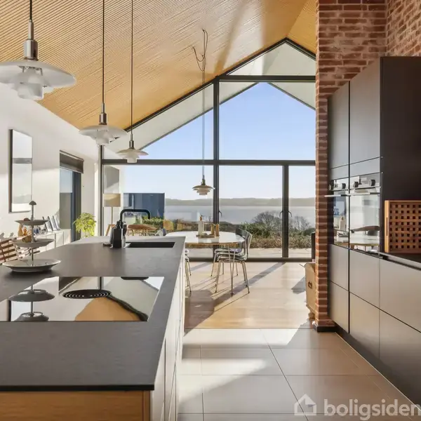 Et moderne køkken med en mørk køkkenø i centrum, omgivet af lyse trælofter og murstensvægge. Store vinduer tillader udsigt til et landskab med træer og en flod.