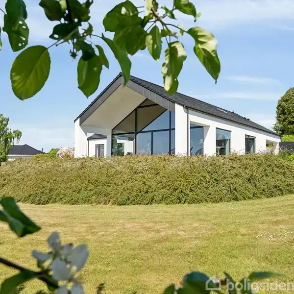 En moderne hvid bygning med store vinduer ligger på en frodig, grøn bakketop omgivet af træer, der rammer motivet. Klar himmel i baggrunden.