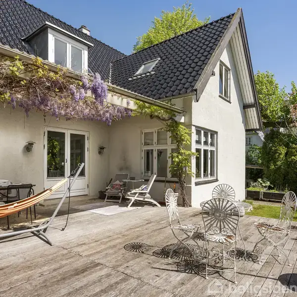 En moderne køkkenindretning med hvide skabe og en ø-bar med barstole. En åben pejs i væggen brænder lyst, og en stor, rund vægur hænger på væggen.