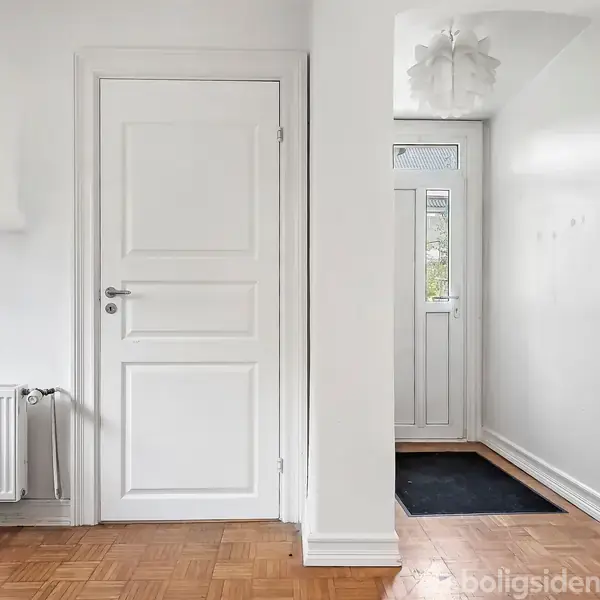 En hvid dør og en radiator står i en lys gang med træparketgulv. En dørmåtte ligger foran en indgangsdør med glasudskæring, der er dekoreret med en loftslampe.