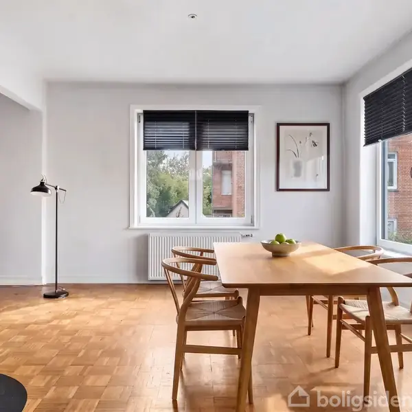 Brændeovn med ild brænder i en lys stue. En sofa står tæt på et stort vindue. Der er et TV på et lavt møbel og lamper i hjørnerne.