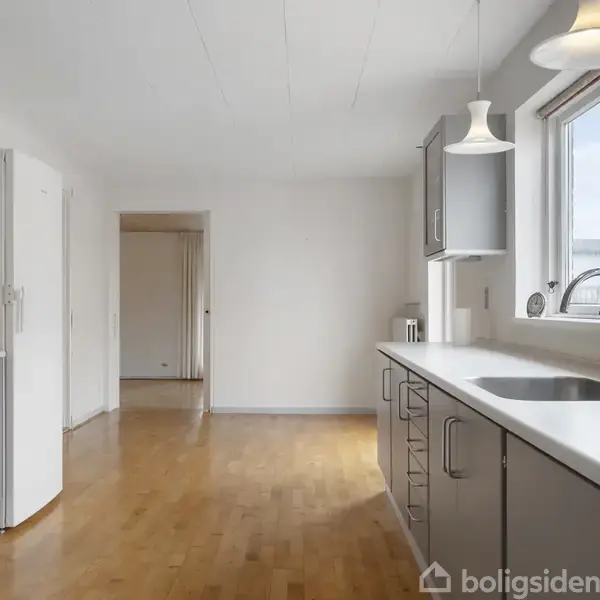 Et køkken med grå skabe og en hvid bordplade, hvor der er monteret en vask. Rummet har trægulv, to vinduer og loftslamper, samt en hvid væg i baggrunden.