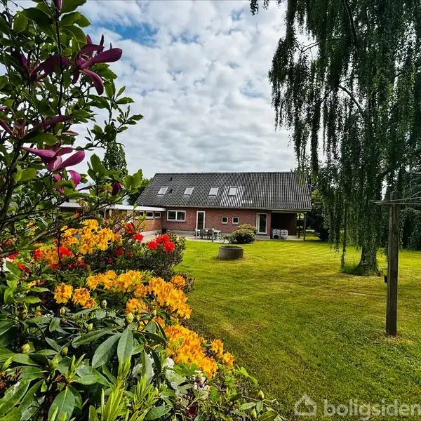 Et rødt hus med flere vinduer og en terrasse står i en stor have med grøn græsplæne. Farverige blomster og træer omkranser området under en skyet himmel.