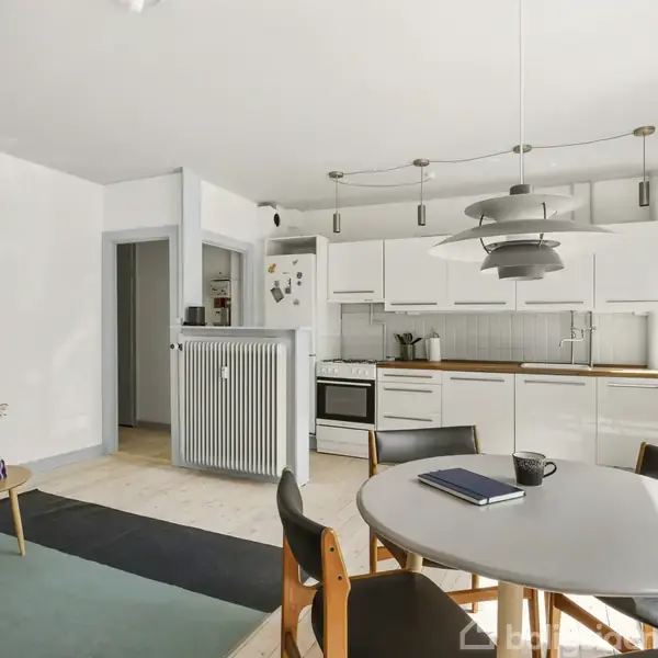 Et lyst køkken-alrum med hvidt køkken, runde spisebord med stole, og en lænestol. Blomster står på et sofabord. En moderne lampe hænger fra loftet. En notesbog ligger på bordet.