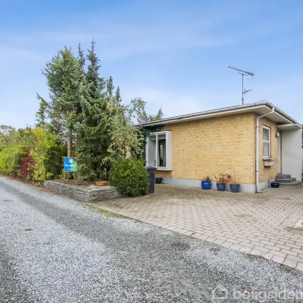 Et gulstenshus med fladt tag står ved siden af en grussti. Foran huset er en brolagt forplads med blomsterkrukker. Omkring er der grønne træer og buske.