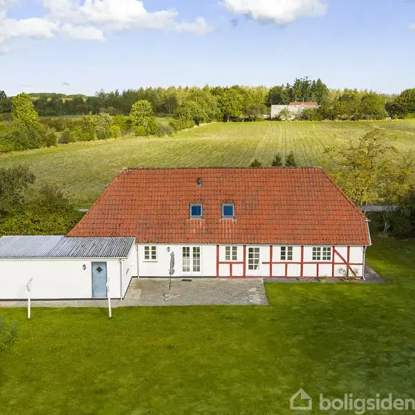 Hvid murstenshus med rødt tegltag står på en grøn græsplæne. I baggrunden ses en mark og træer. Solrigt vejr med få skyer på himlen.