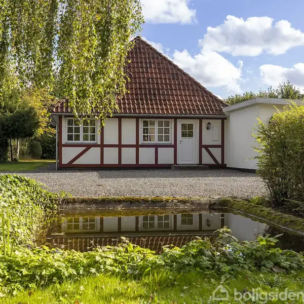 Hus med rødbrunt tag og bindingsværk står foran en grusgård. I forgrunden en lille dam omgivet af grønne planter. Baggrund med blå himmel og træer.