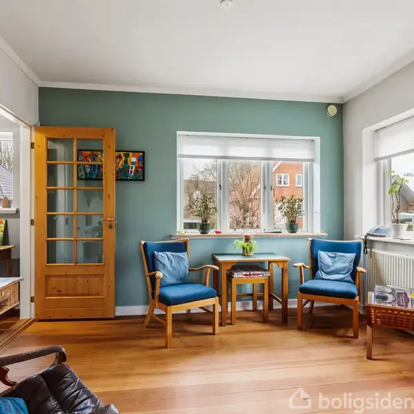 Stue med blåmalet væg indeholder to blå stole og et lille bord ved vindue med udsigt til hus og træer. Trægulv, reol, vægur og grønne planter skaber en lys hjemlig atmosfære.