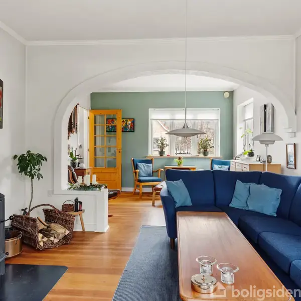Brændeovn opvarmer stue med blå hjørnesofa og træbord. I baggrunden ses spiseplads med træstole, grøn væg og malerier, skønt oplyst af store vinduer.