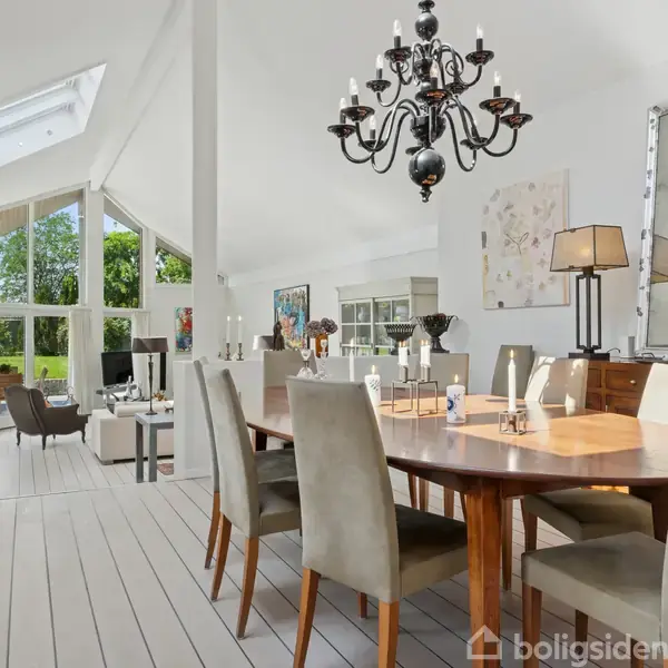 Spisebord med stole i træfyldt stue, dekoreret med en lysekrone og lys. Store vinduer viser en grønt haveområde udenfor. Møbler og vægkunst giver varme og elegance.