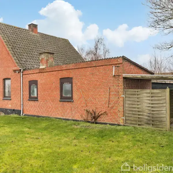 Rødt murstenshus med en skråtag og tre små vinduer, stående på en grøn græsplæne. I baggrunden træer og en blå himmel med skyer.