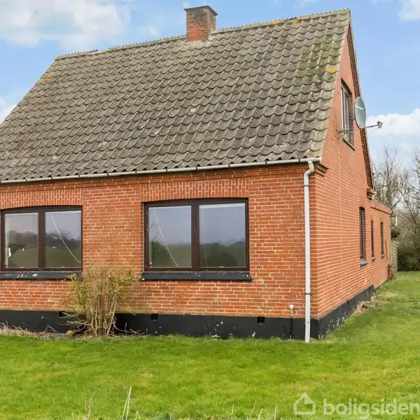 Rødt murstenshus med skråt tag står stille på en grøn græsplæne omgivet af træer og buske. Huset har både vinduer og en parabol antenne på siden.