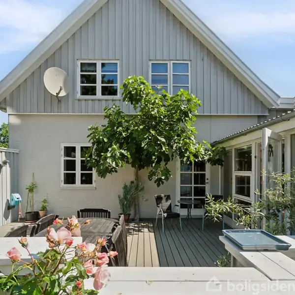 Et hus med gråmalet facade står i en solrig terrassehave. Et træ vokser op ved siden af et bord med stole. Blomster og grønne planter omgiver området.