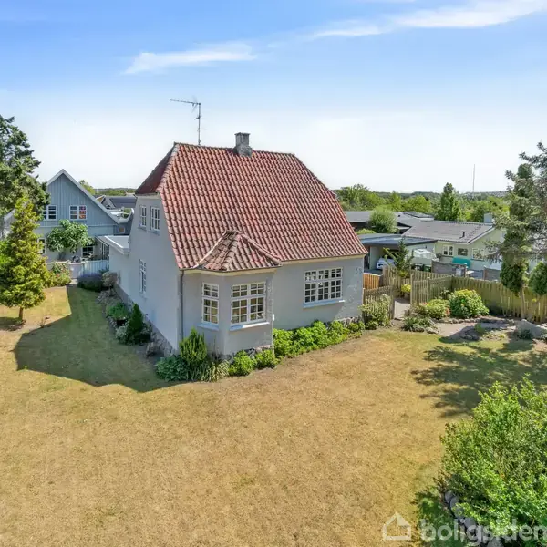 En hvid, to-etagers villa står med en rød tegltag. Den er omgivet af en velholdt have med græsplæne og træer, beliggende i et roligt boligområde.