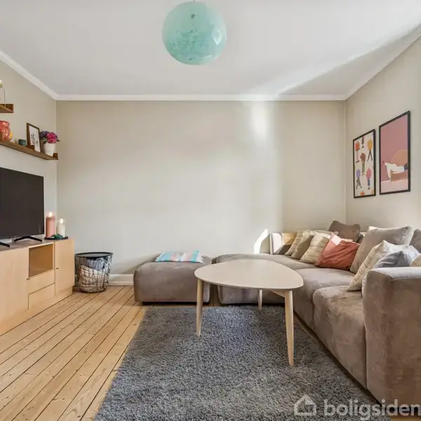 Stor grå sofa med farverige puder står i hjørne, ved siden af et lille rundt bord på lysebrunt trægulv. TV på skænk, hylder og vægkunst i lys stue.