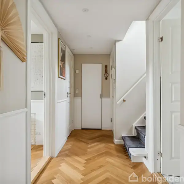 En lys entré med trægulv, hvor en trappe fører opad til højre. En dør for enden af gangen og flere værelser på hver side. En vægdekoration hænger til venstre.