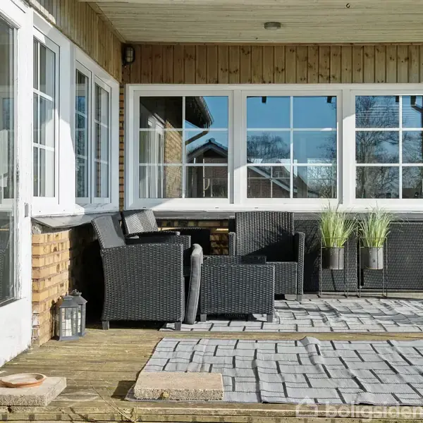Terrassemøbler står på en flisebelagt terrasse foran et hus med gule mursten. Rattan-stole og bord er placeret under store vinduer. To krukker med græsplanter pynter området.