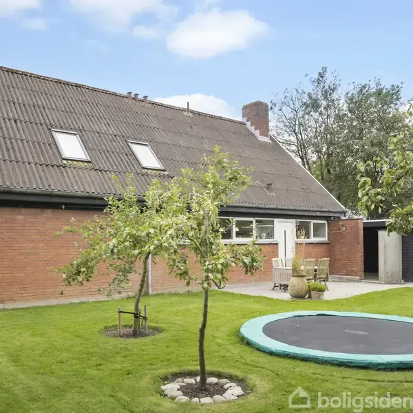 En stor murstensvilla med skråtag står omgivet af en grøn have. En trampolin er nedgravet i græsplænen foran huset, og der er et lille træ ved siden af.
