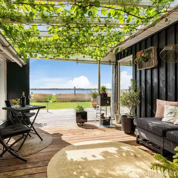 Overdækket terrasse med grønne planter på træværk. Sorte møbler står på træplanker. Udsigt til en solrig have og en sø i det fjerne. Ingen tekst til stede.
