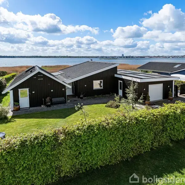 Sort træhus med gråtag står stille i velplejet have, omgivet af grøn hæk. I baggrunden ses kystlinje og vand under en delvist overskyet himmel.