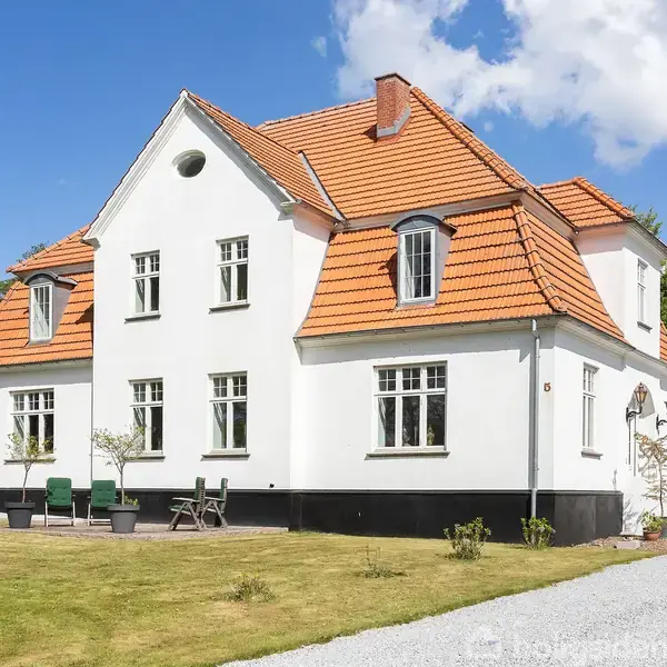 Et hvidt hus med rødt tegltag står på en velholdt græsplæne. Foran huset er der et lille terrasseområde med havemøbler. Solrigt vejr med blå himmel og hvide skyer.