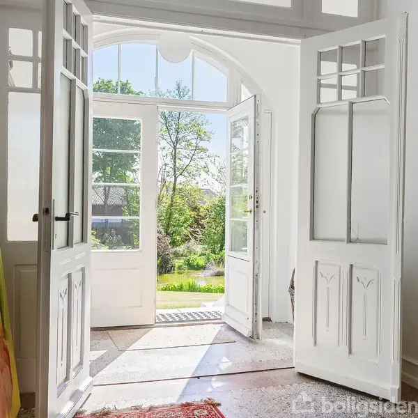 En dobbeltdør står åben mod en solrig have, filtrerer lys ind i en lys indgang. Et stort maleri står ved siden af døren.